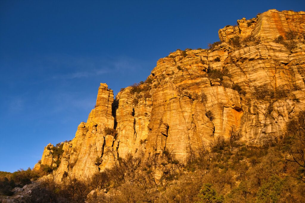 Rugged rock formations with golden sunlight against blue sky and sparse bushes wallpaper background