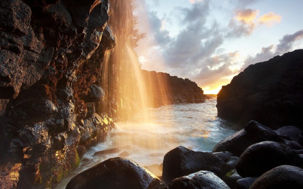 Sunlit waterfall cascading over dark rocks with mist and warm golden hues in tranquil nature Wallpaper