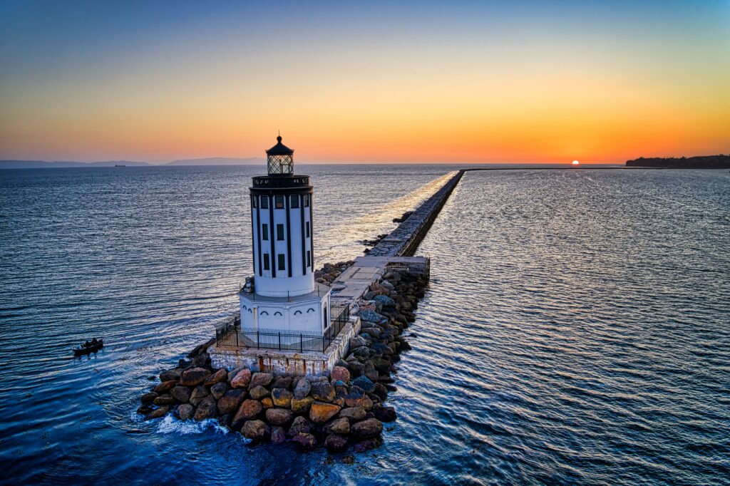 Classic lighthouse on rocky shore with stone pier at sunrise over calm ocean wallpaper background