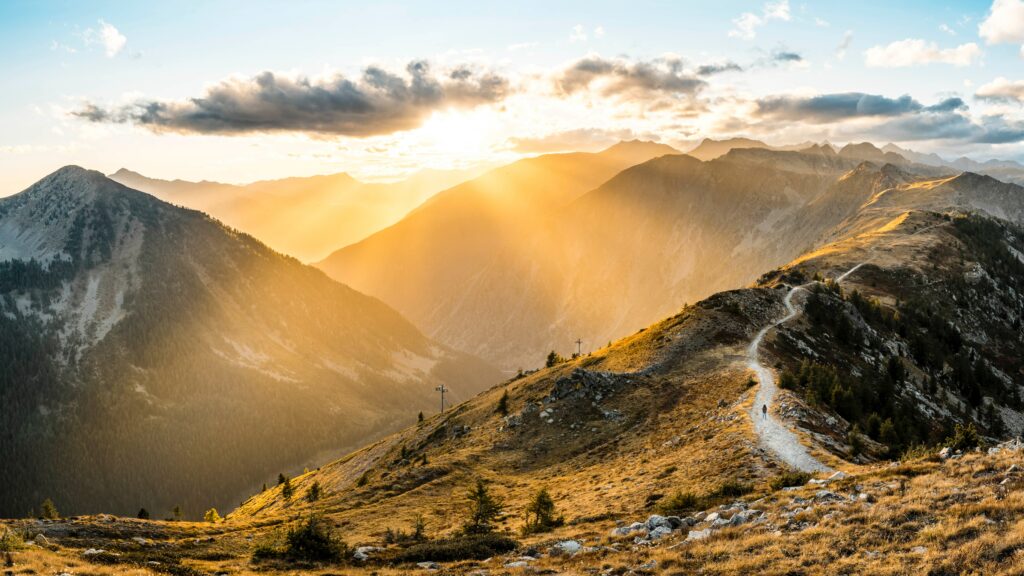 Winding mountain path at sunrise with golden sunlight and dramatic clouds in rugged terrain Wallpaper