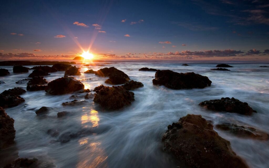 Golden sunlight over rugged coastal rocks with gentle sea foam under vibrant orange blue sky Wallpaper