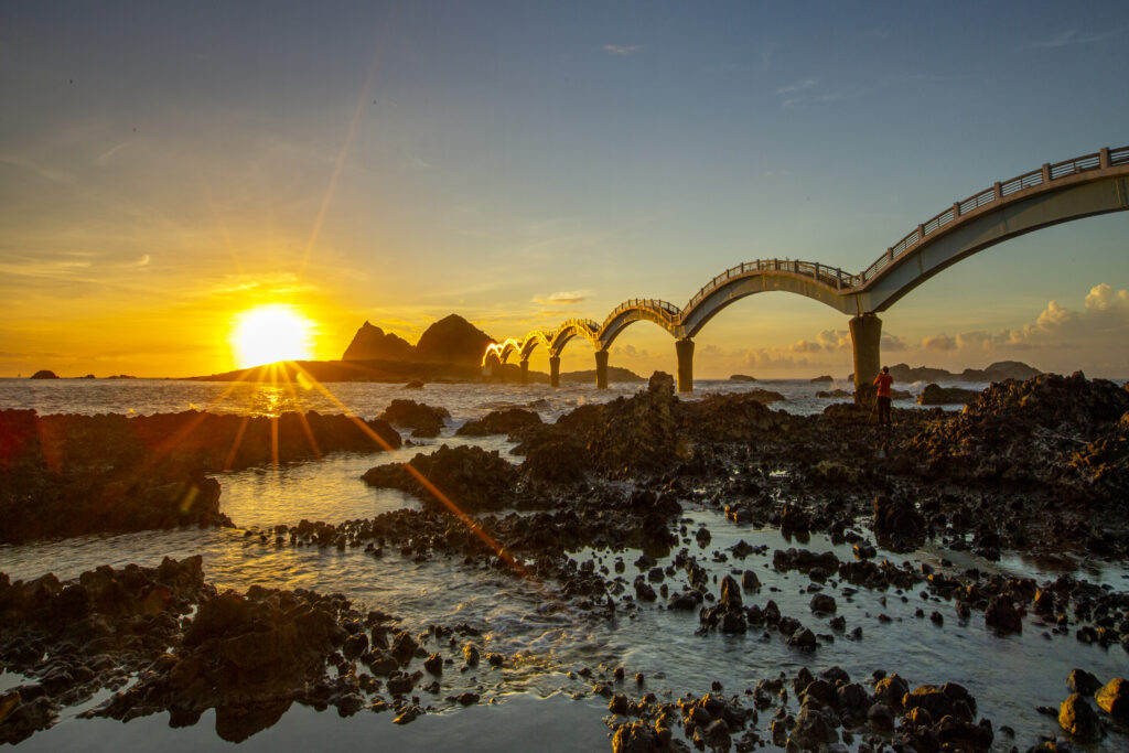 Golden sunrise over calm sea rugged rocks and elegant arched bridge nature architecture harmony Wallpaper