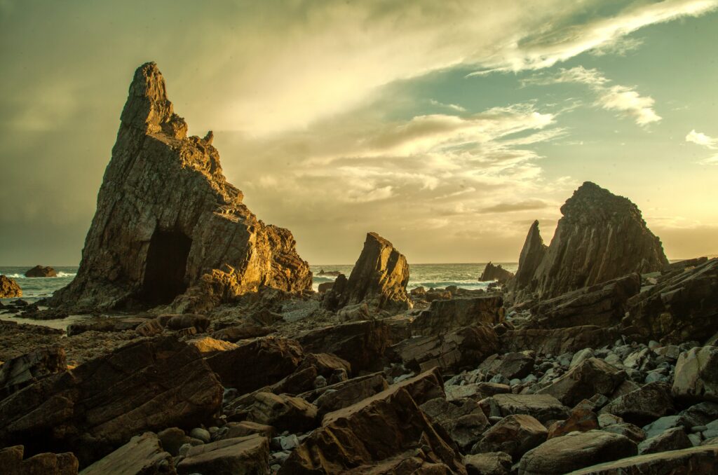 Golden sunset over dramatic jagged rock coastline with scattered stones and soft cloud sky Wallpaper