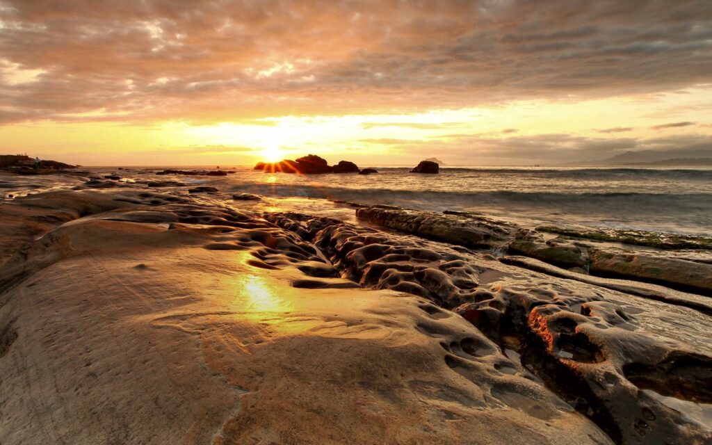 Sunset coastal wallpaper with golden light on rocky shoreline and dramatic clouds over ocean