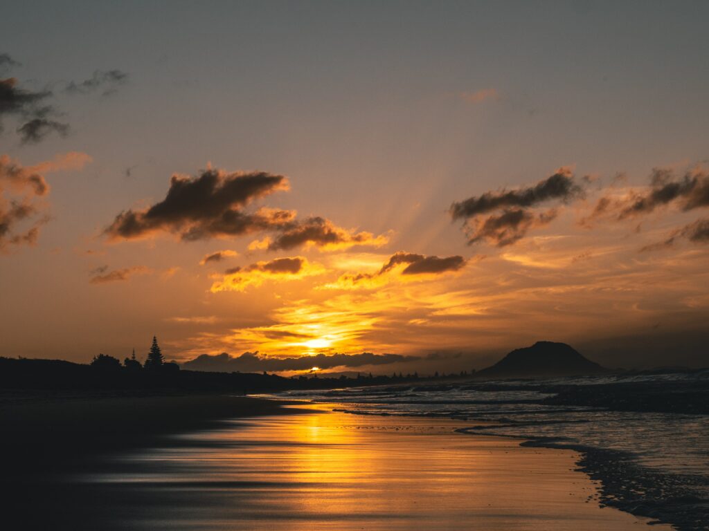 Sunset over calm sea and wet sand reflecting golden amber hues with soft clouds and distant hills Wallpaper