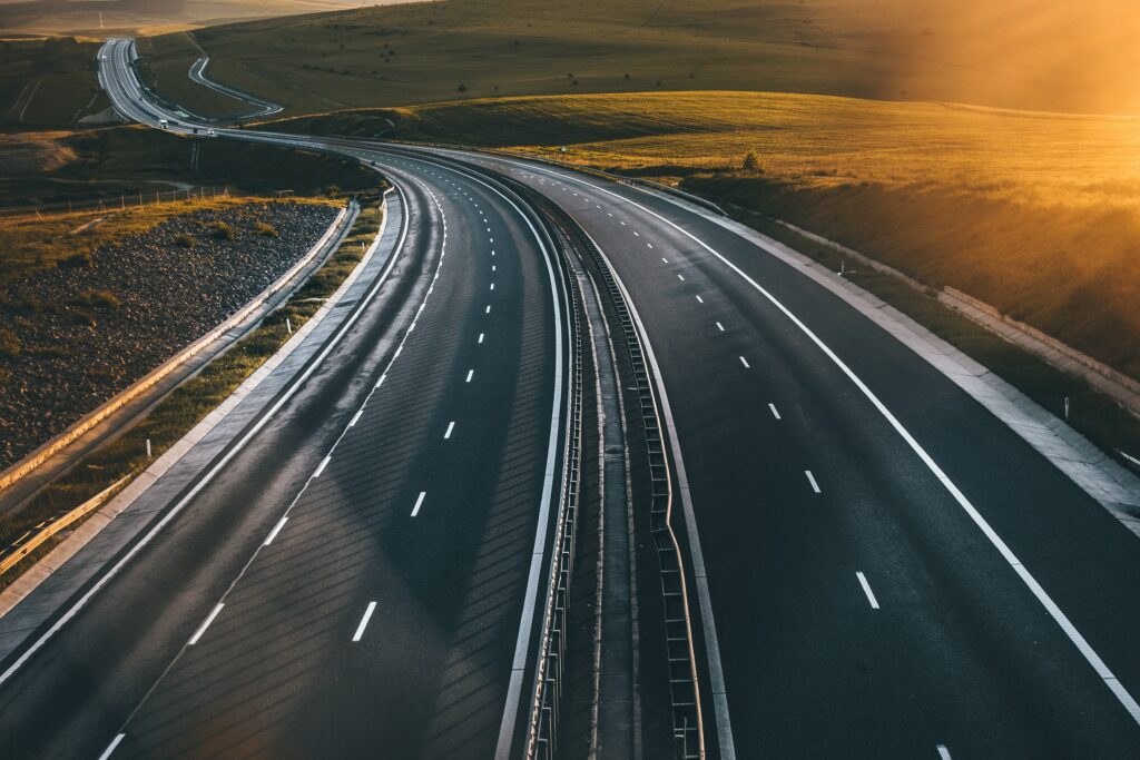 Smooth asphalt highway with white markings under golden sunset bordered by hills and fields Wallpaper
