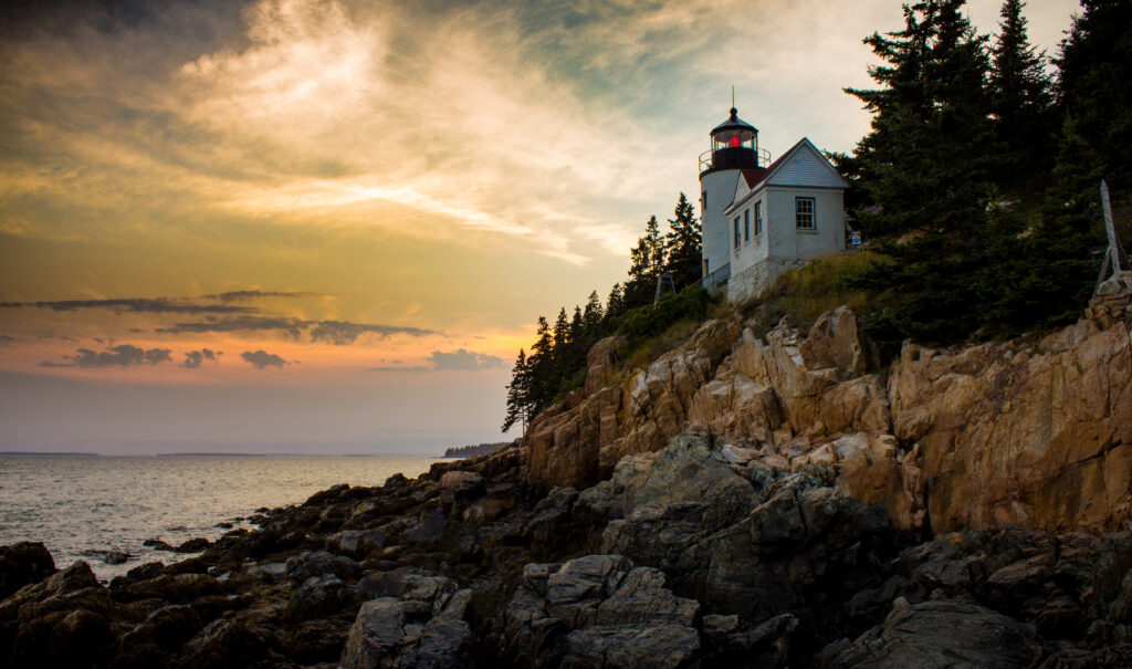 Lighthouse on rugged cliffs at sunset with evergreen trees waves and dramatic coastal sky wallpaper