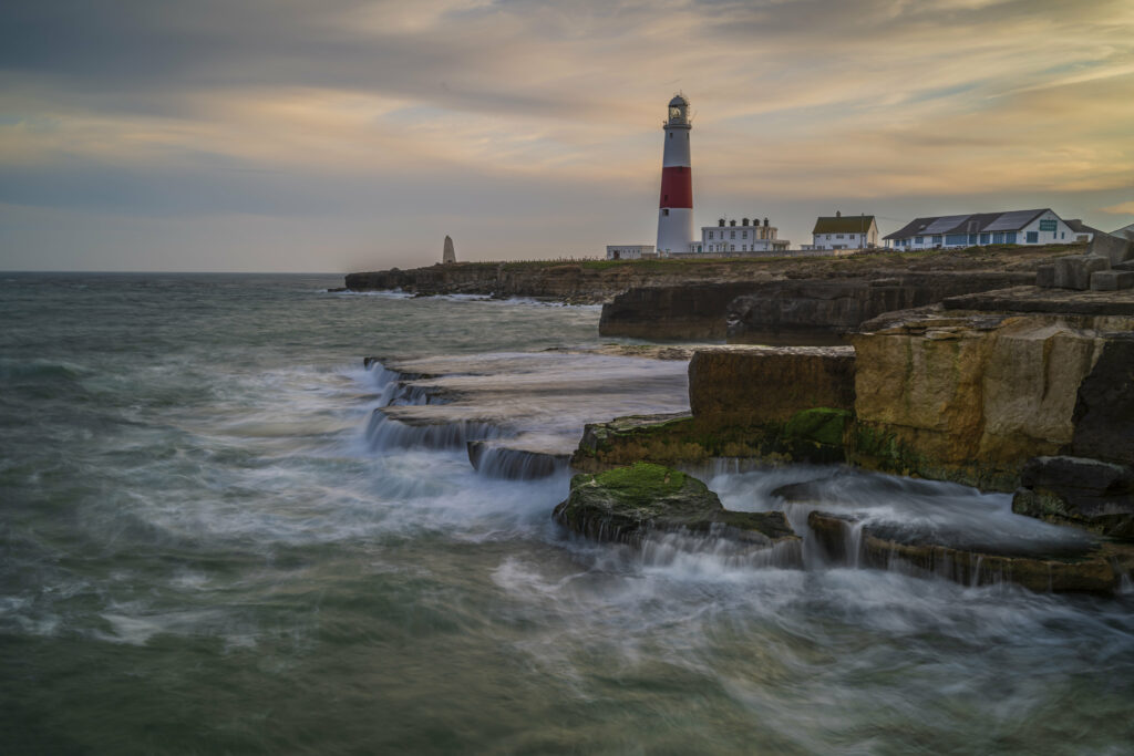 Lighthouse on rocky coast at sunset with crashing waves serene coastal landscape wallpaper background
