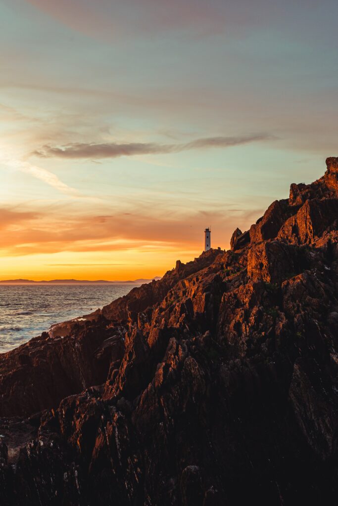 Solitary lighthouse on rugged cliffs at sunset with warm golden sky coastal wallpaper background