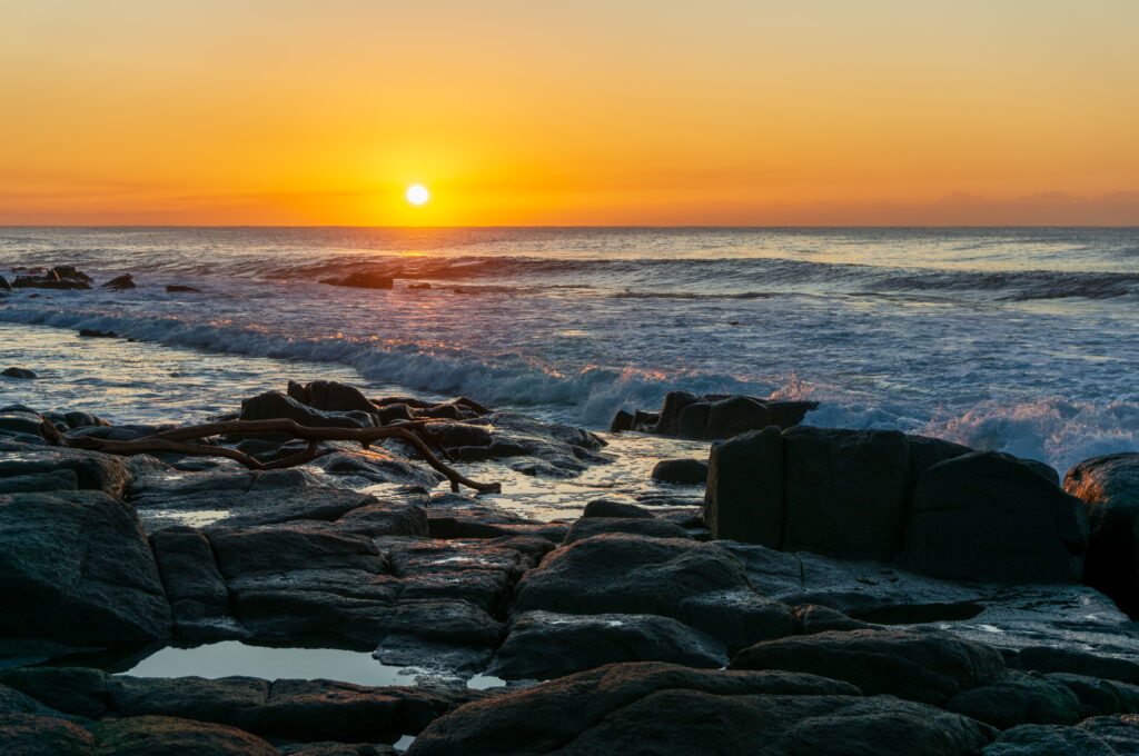 Golden sunset lighting dark coastal stones with gentle waves creating a tranquil seaside scene Wallpaper