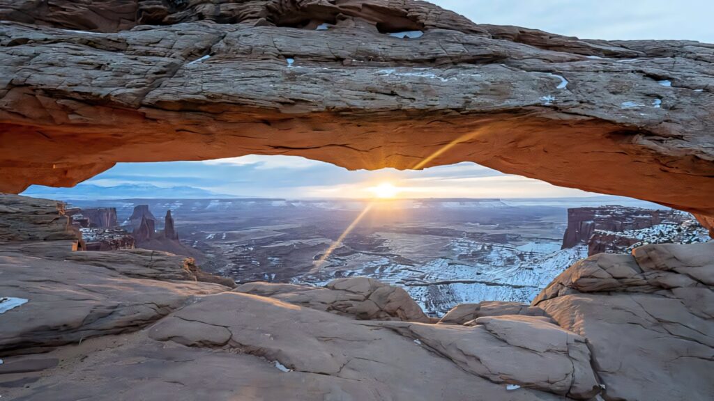 Breathtaking sunset through Delicate Arch casting warm glow on red rocks and snowy valleys Wallpaper