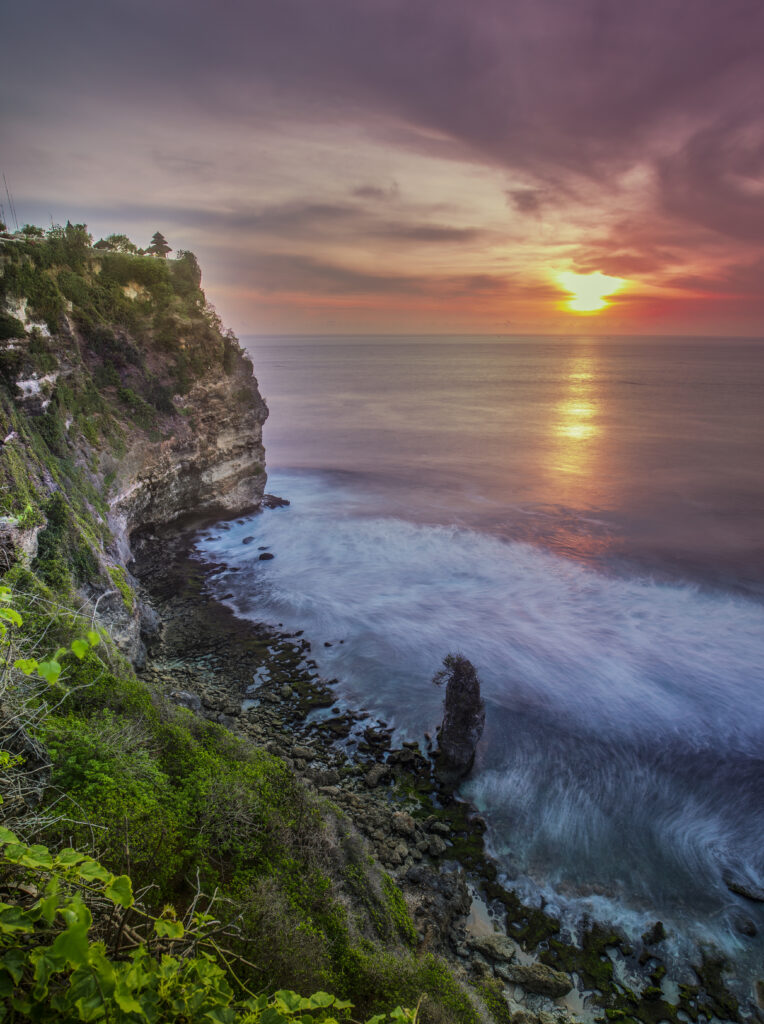 Dramatic rock cliff above serene shore with golden sunset and gentle waves in vibrant colors Wallpaper