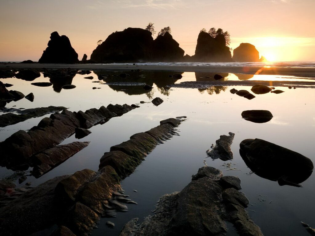 Dramatic rock formations reflect golden sunset hues over tranquil water with a glowing horizon Wallpaper
