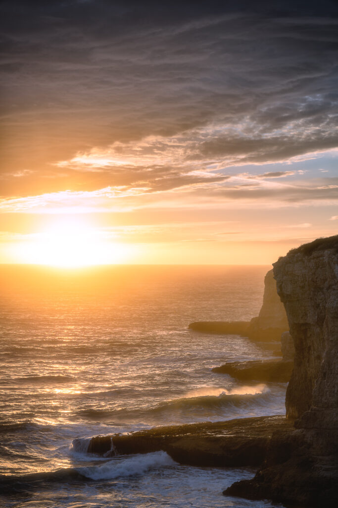 Sunset over rocky coastline with golden sunlight reflecting on waves and dramatic clouds wallpaper