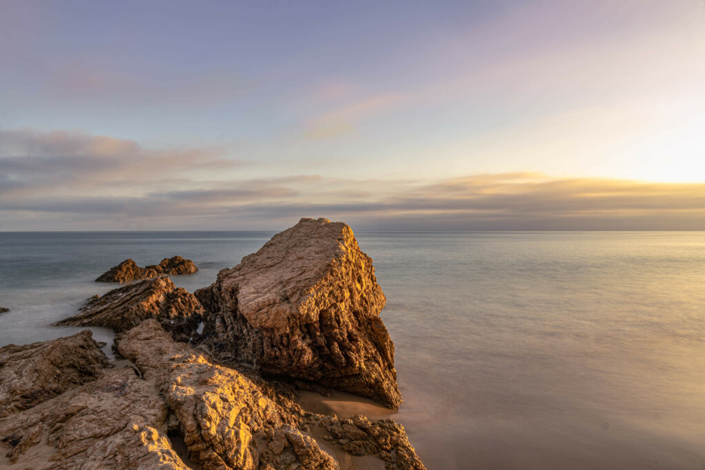 Golden sunset over rugged rocks and calm sea waters reflecting warm hues in tranquil sky Wallpaper
