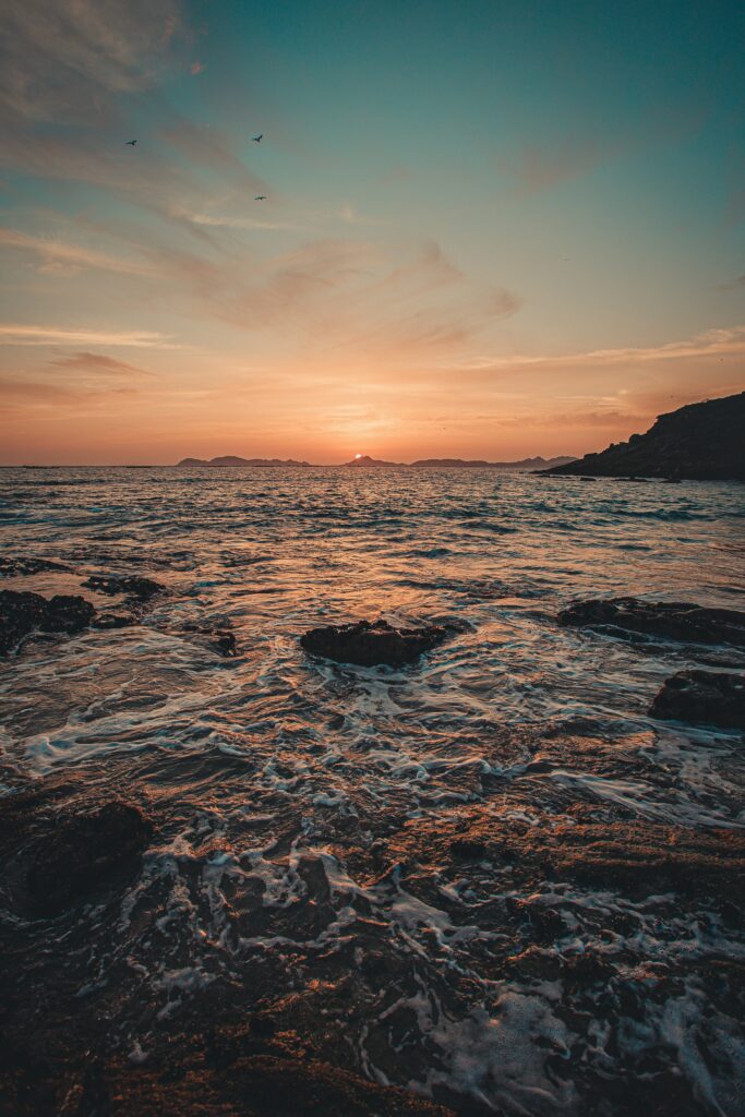 Sunset over sea with golden orange reflection waves rocky shoreline soft clouds and hills wallpaper