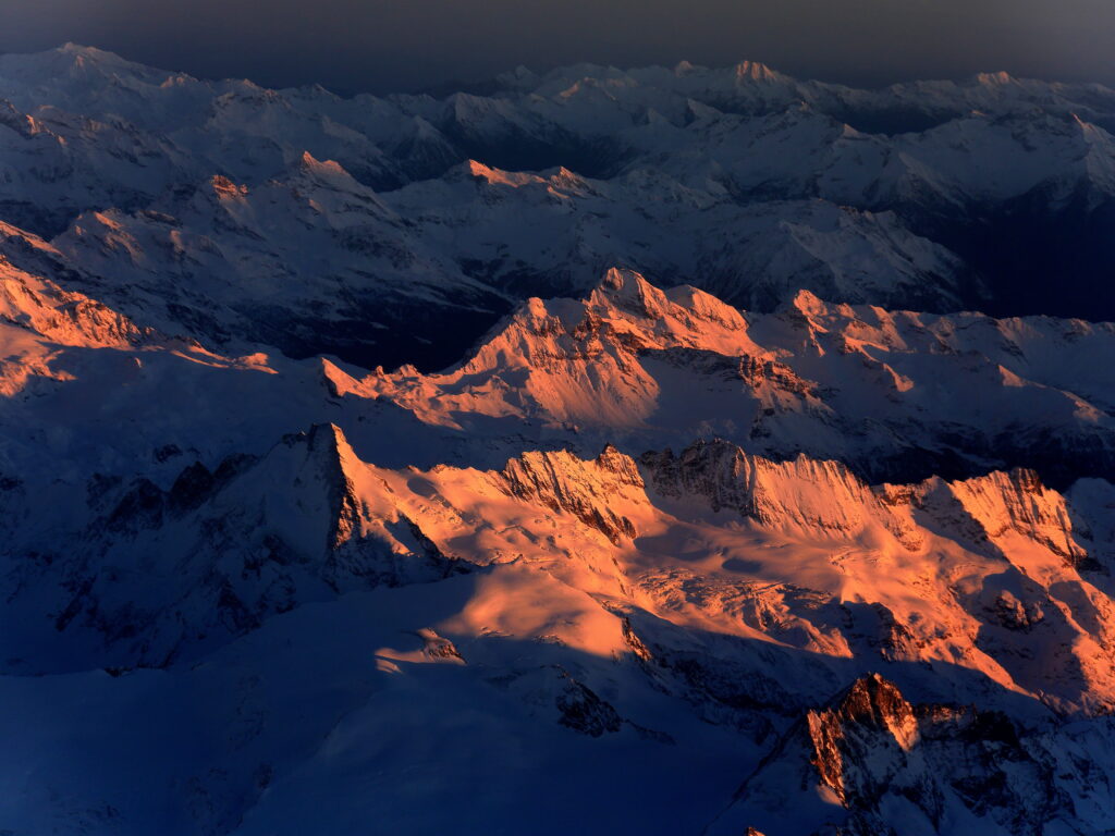 Aerial view of snow-covered mountains at dusk with golden sunset light and dramatic shadows Wallpaper