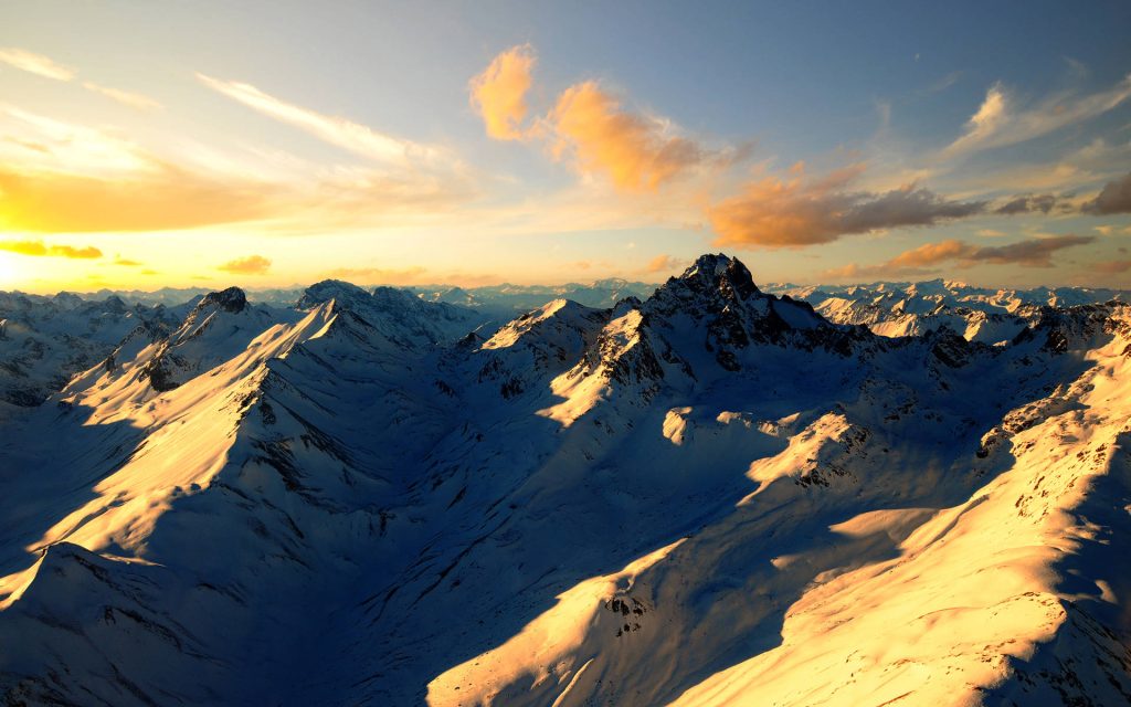 Snowy mountain range at sunset with golden light highlighting jagged peaks and deep valleys Wallpaper