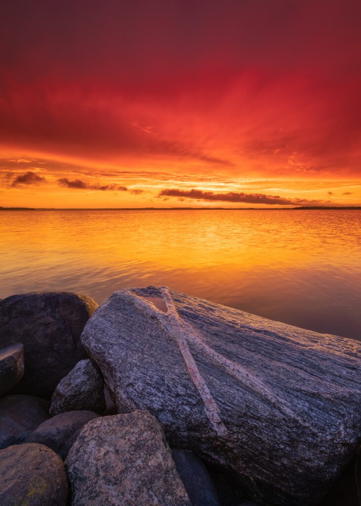 Large textured stones foreground with golden sunset over calm water horizon vibrant red sky wallpaper