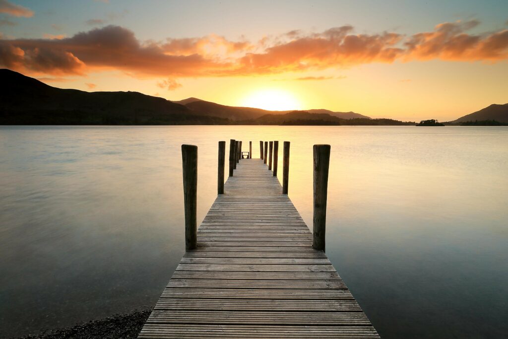 Wooden pier over tranquil lake at sunset with golden hues calm water and gentle hills landscape Wallpaper