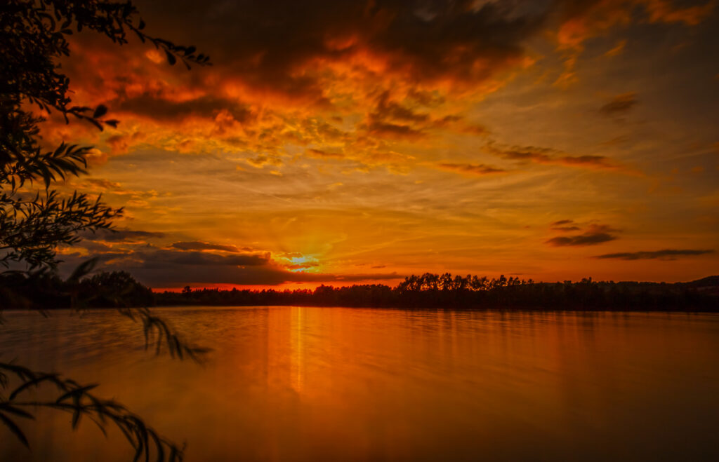 Sunset wallpaper with vivid orange sky reflecting on river framed by silhouetted trees and clouds