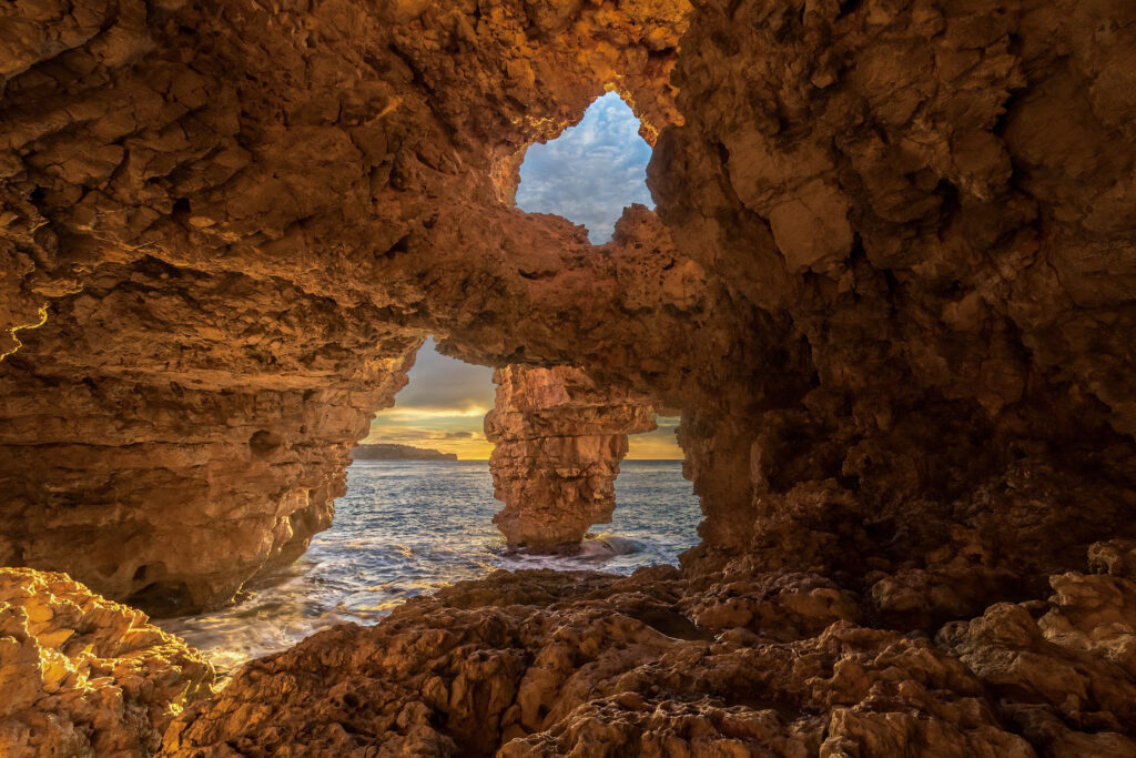 Natural cave opening to sea at golden sunset with rugged stones and warm earthy tones wallpaper