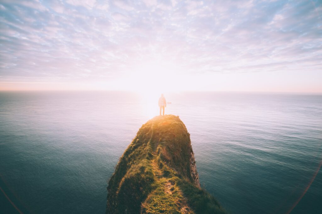 Silhouette on rugged cliff overlooking calm sea at golden sunset with soft sunlight and clouds Wallpaper