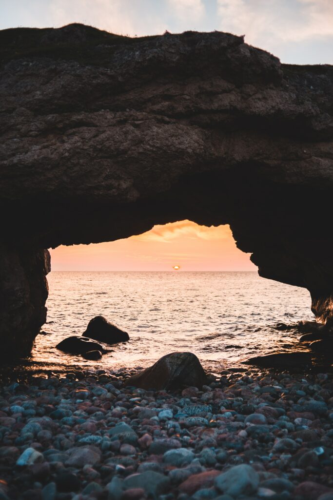 Natural stone arch framing golden sunset over calm sea with smooth pebbles and weathered rocks Wallpaper