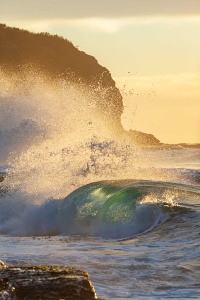 Powerful wave crashes rugged coastline at sunset with golden light and vibrant sea spray wallpaper