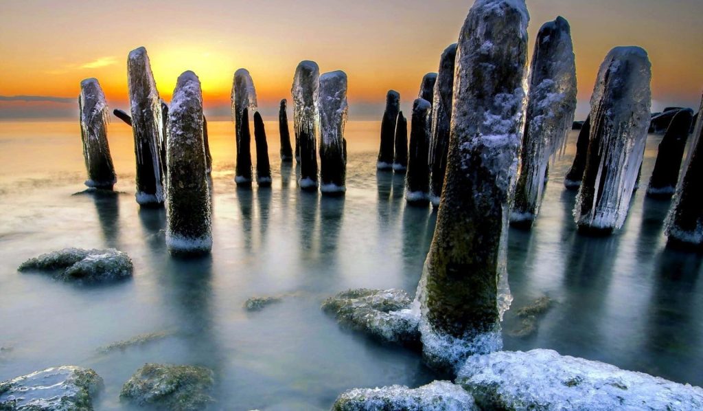 Winter wallpaper with icy wooden posts in calm water at golden sunset shore scene