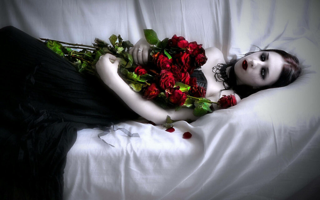 Gothic vampire bride in black lies on white bed with red roses dark makeup vampire wallpaper background