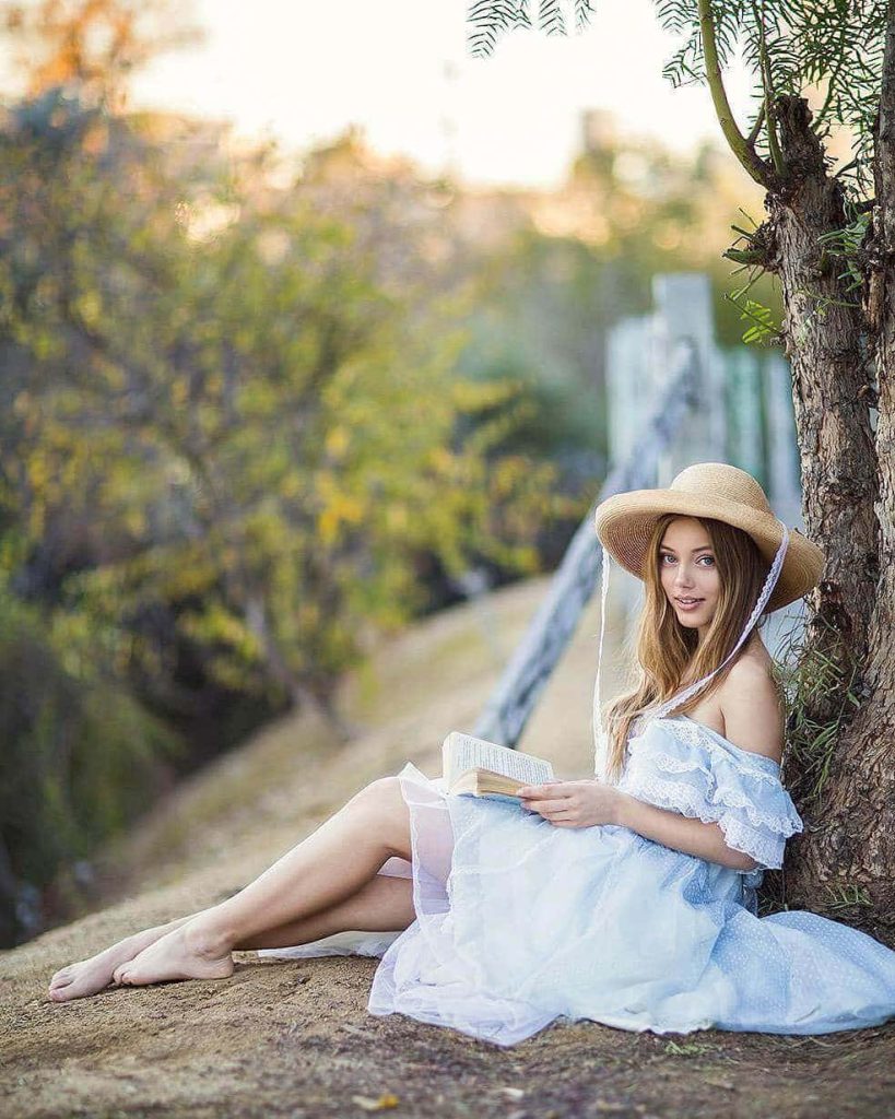 Grace Van Dien in blue ruffled dress and straw hat reading book under tree golden light Wallpaper