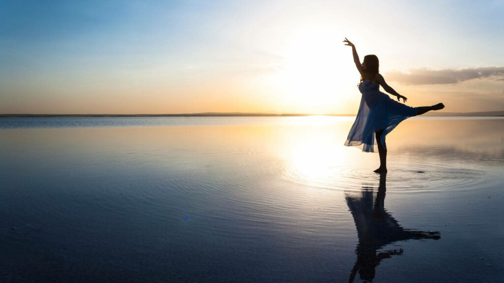 Ballet dancer silhouette in flowing dress at sunset reflected on calm water serene wallpaper