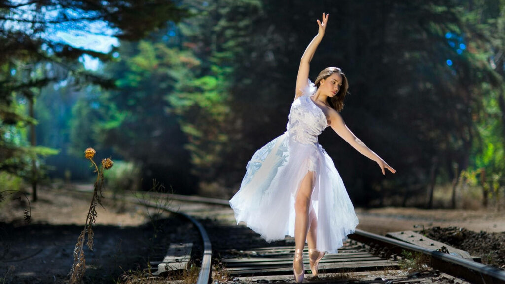 Ballet dancer in flowing white dress performing gracefully on sunlit train track with greenery Wallpaper