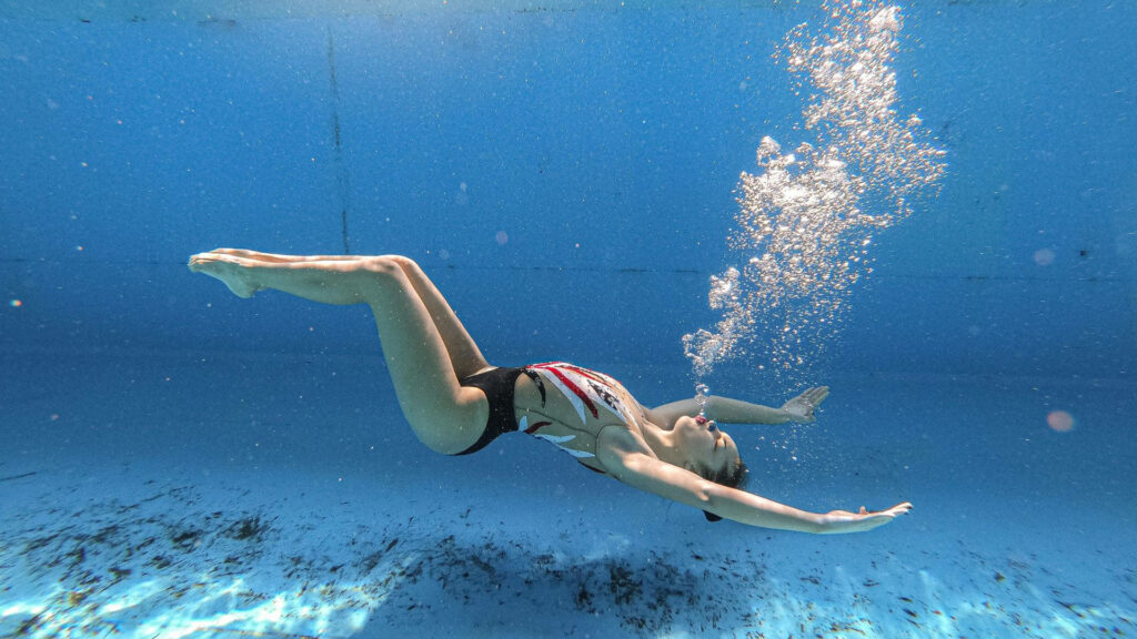 Breathtaking Artistry: A mesmerizing underwater vista of an elegant artistic swimmer gracefully submerging with remarkable form and captivating bubbles Wallpaper