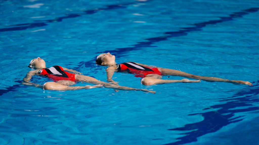 Two artistic swimmers in red swimsuits synchronized floating on backs in clear blue pool wallpaper