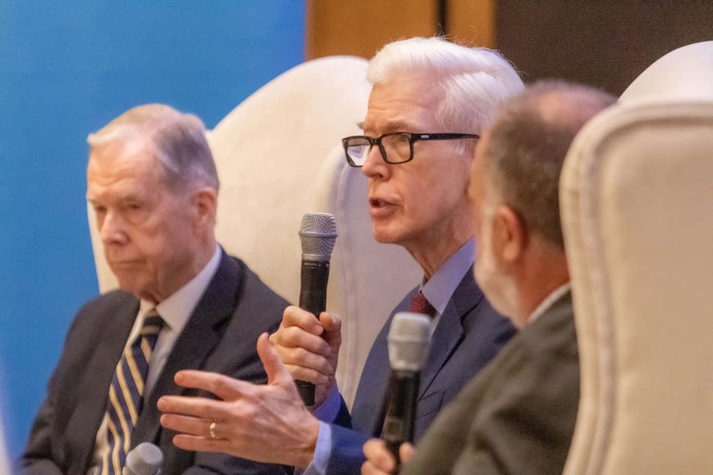 Gray Davis speaking into microphone panel discussion high-backed white chairs formal setting wallpaper