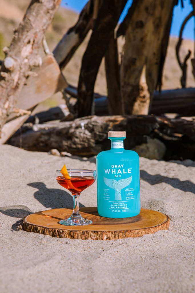 Gray Whale Gin bottle with red cocktail and orange peel on rustic board by sandy driftwood beach Wallpaper