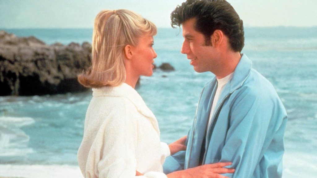 Grease 1978 beach scene wallpaper with Danny Zuko and Sandy Olsson iconic romantic gaze