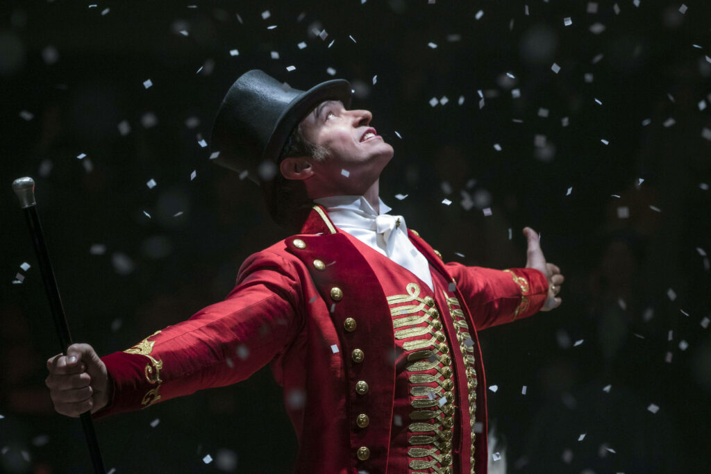 The Greatest Showman star in red ringmaster costume with arms open under confetti wallpaper