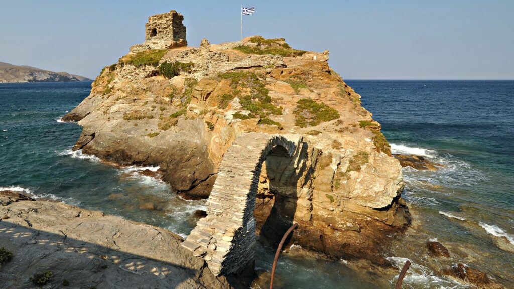 Greek rock formation with stone arch bridge ancient ruins and flag above clear blue sea sky Wallpaper