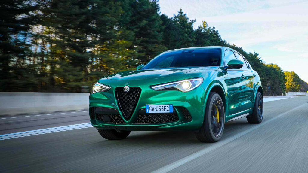 Green Alfa Romeo Stelvio driving on open road with lush trees and clear blue sky background Wallpaper