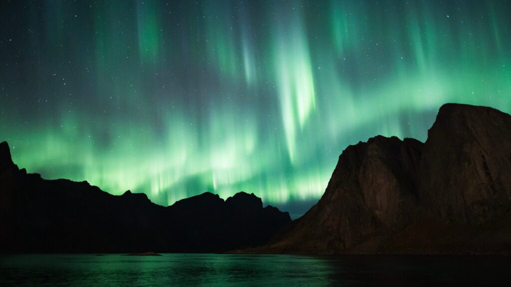 Vivid green northern lights over rugged mountains reflecting on a calm lake at night wallpaper
