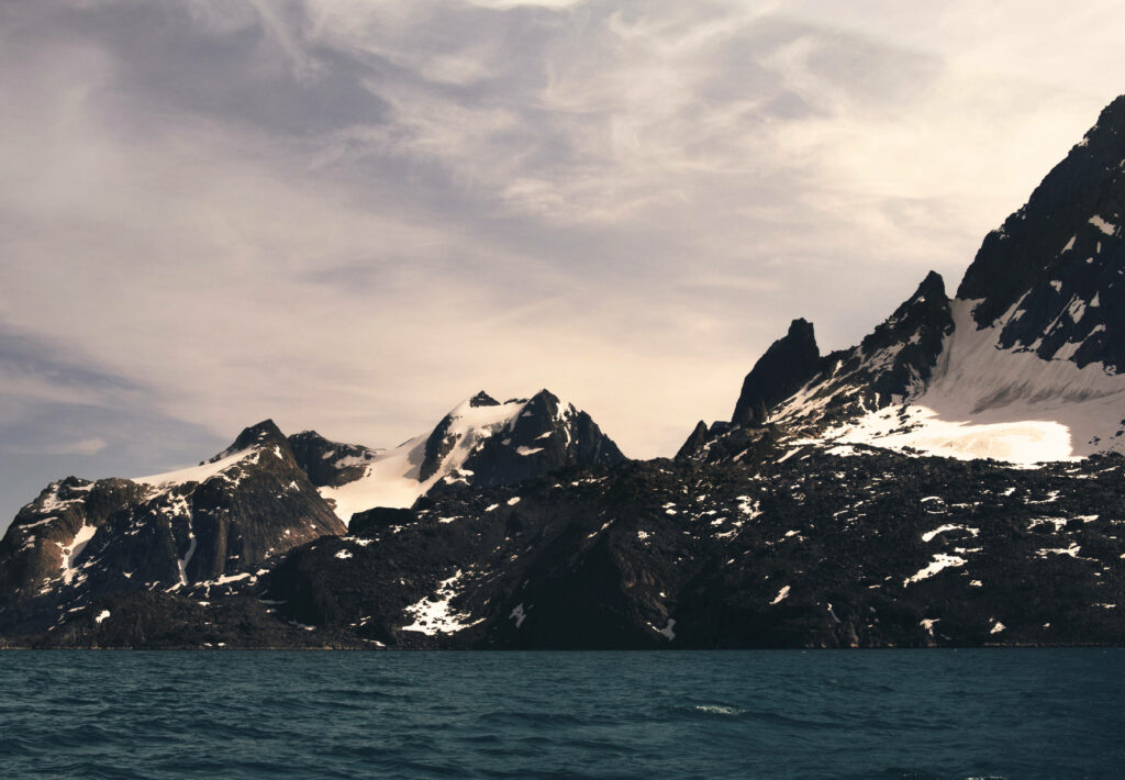Snow-capped Greenland Alps towering over deep blue Arctic waters under a dramatic sky wallpaper