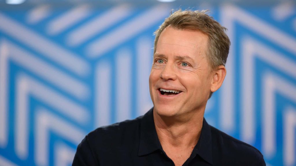 Greg Kinnear smiling in dark polo with modern blue white geometric wallpaper background
