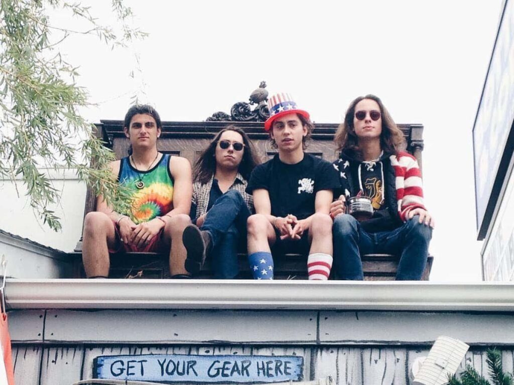 Greta Van Fleet band sitting on rooftop ledge under Get Your Gear Here sign in natural light Wallpaper