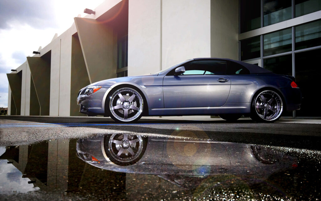 Grey convertible with professional detailing reflecting in water puddle outside modern building wallpaper