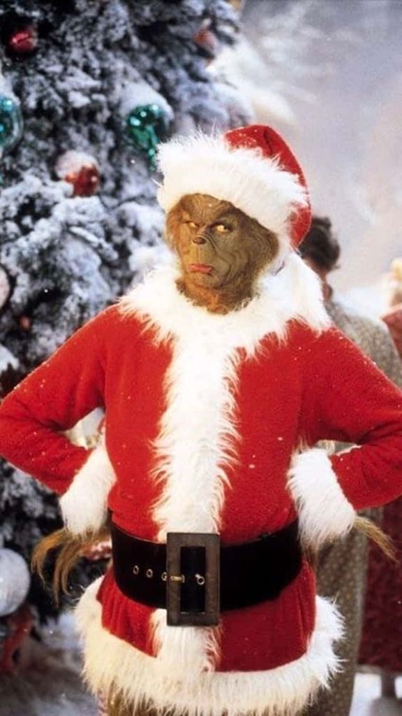Jim Carrey as Grinch in Santa suit by snow-covered Christmas tree festive wallpaper background