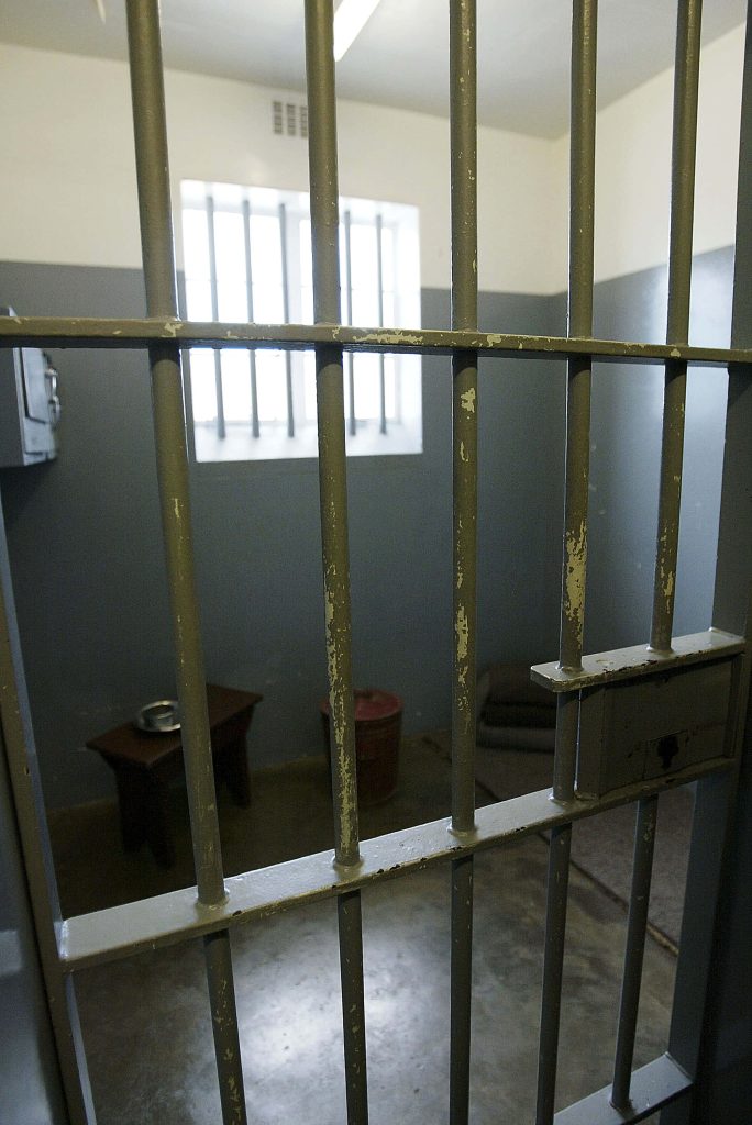 Dimly lit prison cell with weathered bars wooden bench metal bucket and small window background Wallpaper