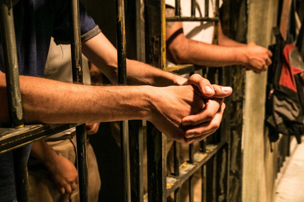 Clasped inmate hands behind heavy jail bars with dramatic warm lighting prison wallpaper background